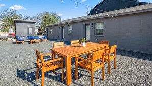 Un bâtiment avec une terrasse en gravier, une table et des chaises en bois, un petit abri de jardin et une voiture au fond.