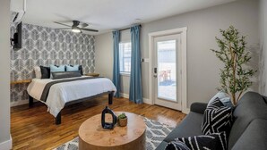 Une chambre à coucher avec un lit, un ventilateur de plafond, une fenêtre avec des rideaux, une plante et une vue sur l’extérieur.