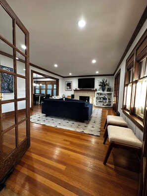 A spacious living room with a dark blue sofa, wooden flooring, a fireplace, and a television mounted on the wall. - Experience Coast Guard Festival Like a Local in a Charming Downtown Cottage (Grand Haven)