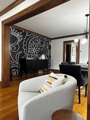 Un salon moderne avec un mur orné d’un motif noir et blanc, une table à manger en bois foncé et un canapé gris agrémenté de coussins à motifs.