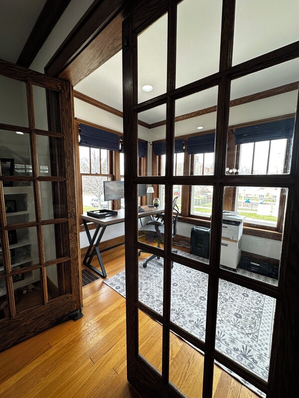 A room with a wooden floor, a desk with a chair, a printer, and a window with a view of the outside. - Experience Coast Guard Festival Like a Local in a Charming Downtown Cottage (Grand Haven)