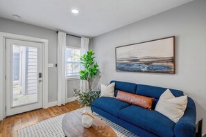 Ein Wohnzimmer mit blauem Sofa, weißer Vase mit Pflanze, einem Bild an der Wand und einer Tür mit Blick.