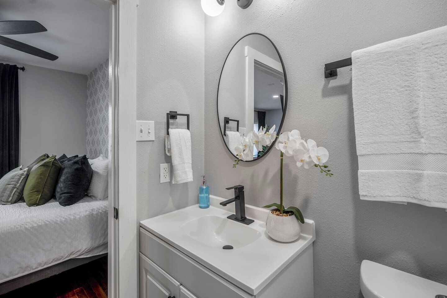 A modern bathroom with a white sink, a mirror, a toilet, and a bed with pillows.