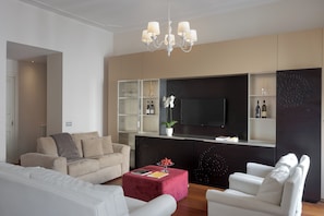 Ein modernes Wohnzimmer mit einem beigefarbenen Sofa, weißen Sessel, einem roten Hocker, einer Chandelier, einem Flachbildfernseher und eingelassenen Regalen mit Dekorationselementen.