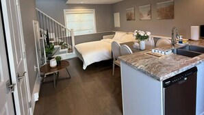 Quarto de hotel moderno com cama, um pequeno espaço de estar, cozinha equipada com lava-louças e escada que leva a um andar superior.