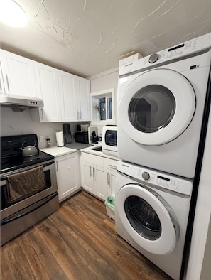 A compact kitchen with white cabinets, a microwave, a kettle, and a stacked washer-dryer unit. - Cozy 2BR Suite · Near Crescent Beach & Trails (Surrey)