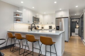 Une cuisine moderne avec des meubles blancs, un réfrigérateur en acier inoxydable, un four à micro-ondes encastré et un coin bar avec des tabourets.