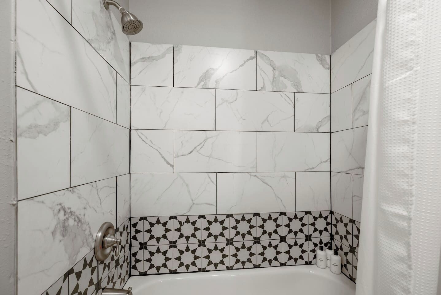 Ein Badezimmer mit weißen Marmorfliesen, einer geometrischen Bordüre und einer Duschbrause.
