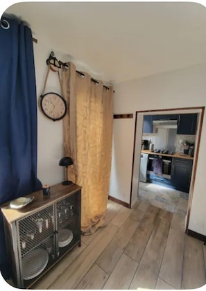 Ein Zimmer mit Holzboden, einem Metallschrank, einer Wanduhr und einer durchgehende Tür erkennbaren Küche.