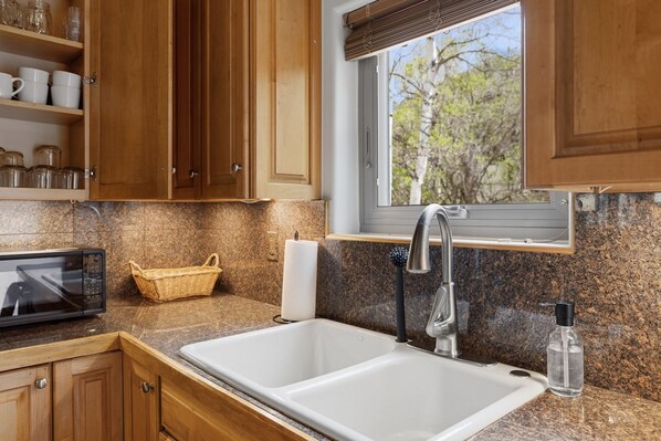 A kitchen with wooden cabinets, a granite countertop, a double sink, a microwave, a basket, and a window with a view of trees. - Cozy Duplex in West Vail – Near Vail Village (Vail)