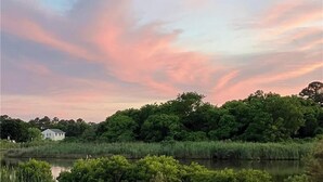 Ett fridfullt landskap med ett hus nära en vattenyta, omgivet av frodig grönska och en färgstark himmel.