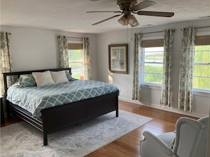 Ein Schlafzimmer mit Bett, Deckenventilator, Fenster mit Vorhängen und einem gerahmten Bild an der Wand.