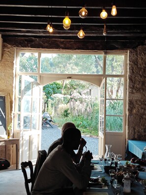 Un coin repas avec une table dressée pour deux personnes, un mur en pierre et une vue sur un jardin, visible à travers une grande porte ouverte.