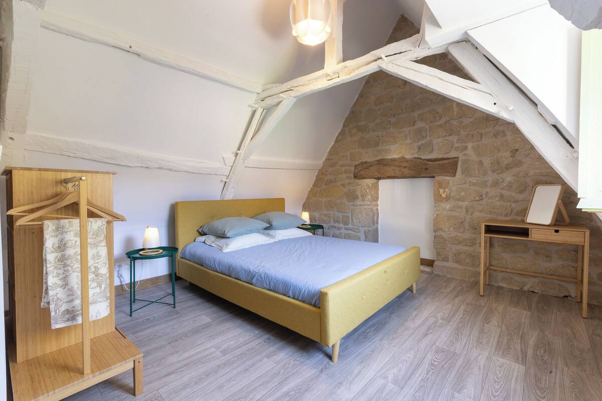 A bedroom with a sloped ceiling, a bed with a wooden headboard, a wooden dresser, a bedside table with a lamp, and a stone wall.