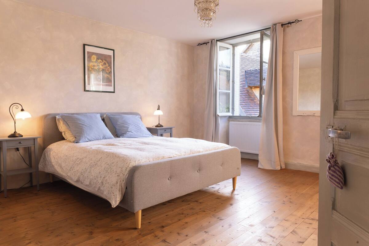 A bedroom with a bed, two bedside tables, a chandelier, a window with curtains, and a radiator.