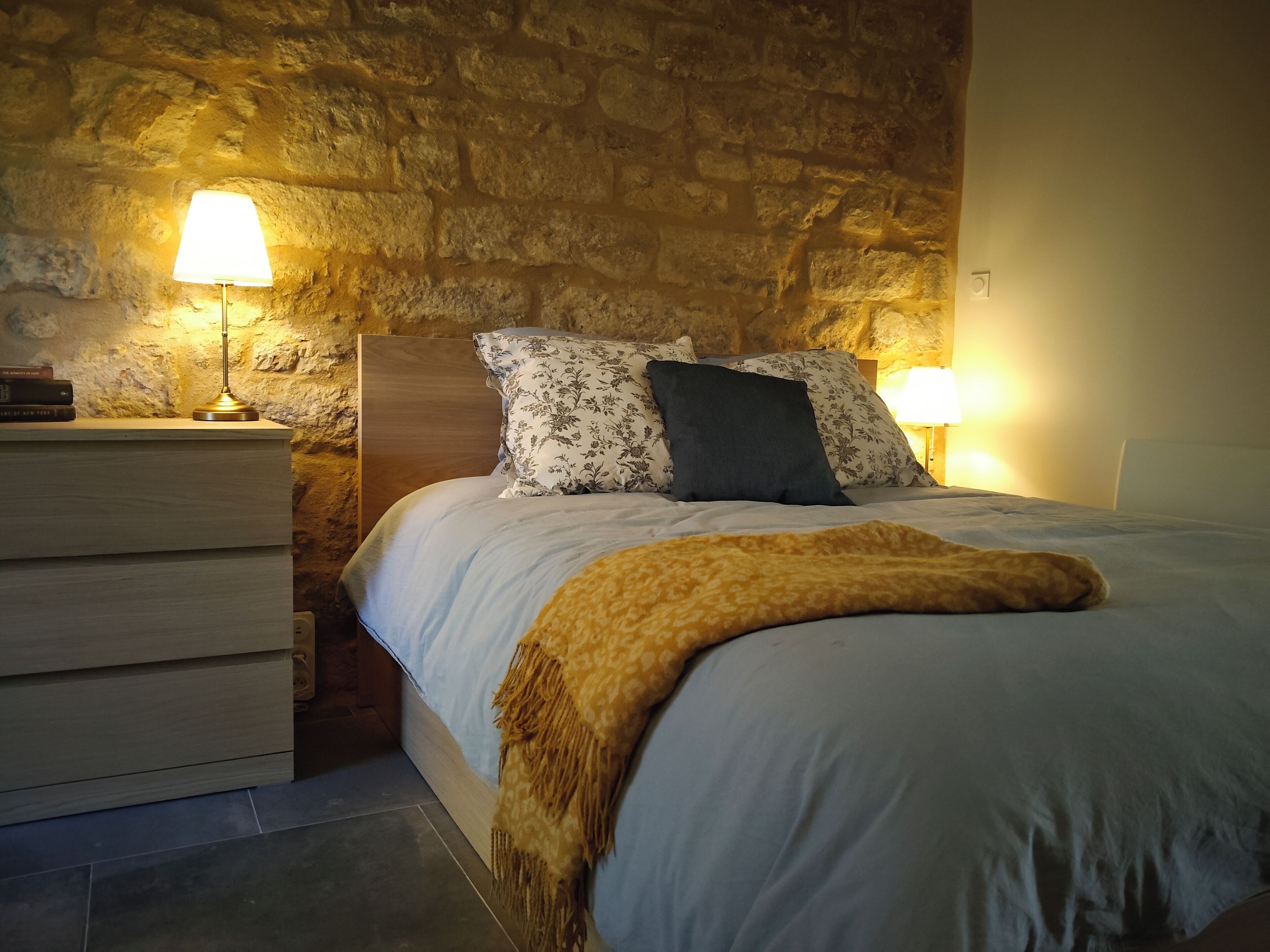 A bedroom with a stone wall, a wooden headboard, a bedside table with a lamp, and a bed with patterned pillows.