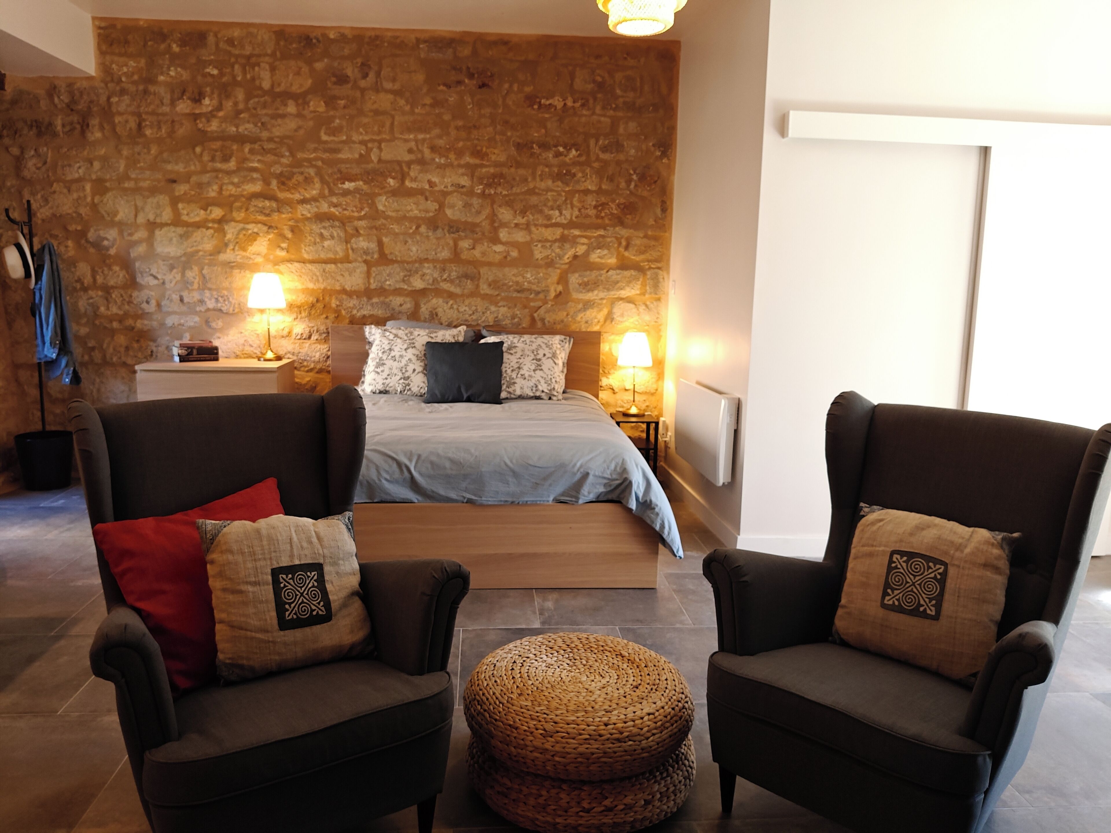 Ein Schlafzimmer mit Steinwand, einem Bett mit blauer Bettdecke, zwei Sesseln mit dekorativen Kissen und einem gewebten Hocker.