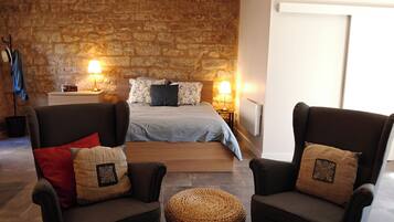 Ein Schlafzimmer mit Steinwand, einem Bett mit blauer Bettdecke, zwei Sesseln mit dekorativen Kissen und einem gewebten Hocker.