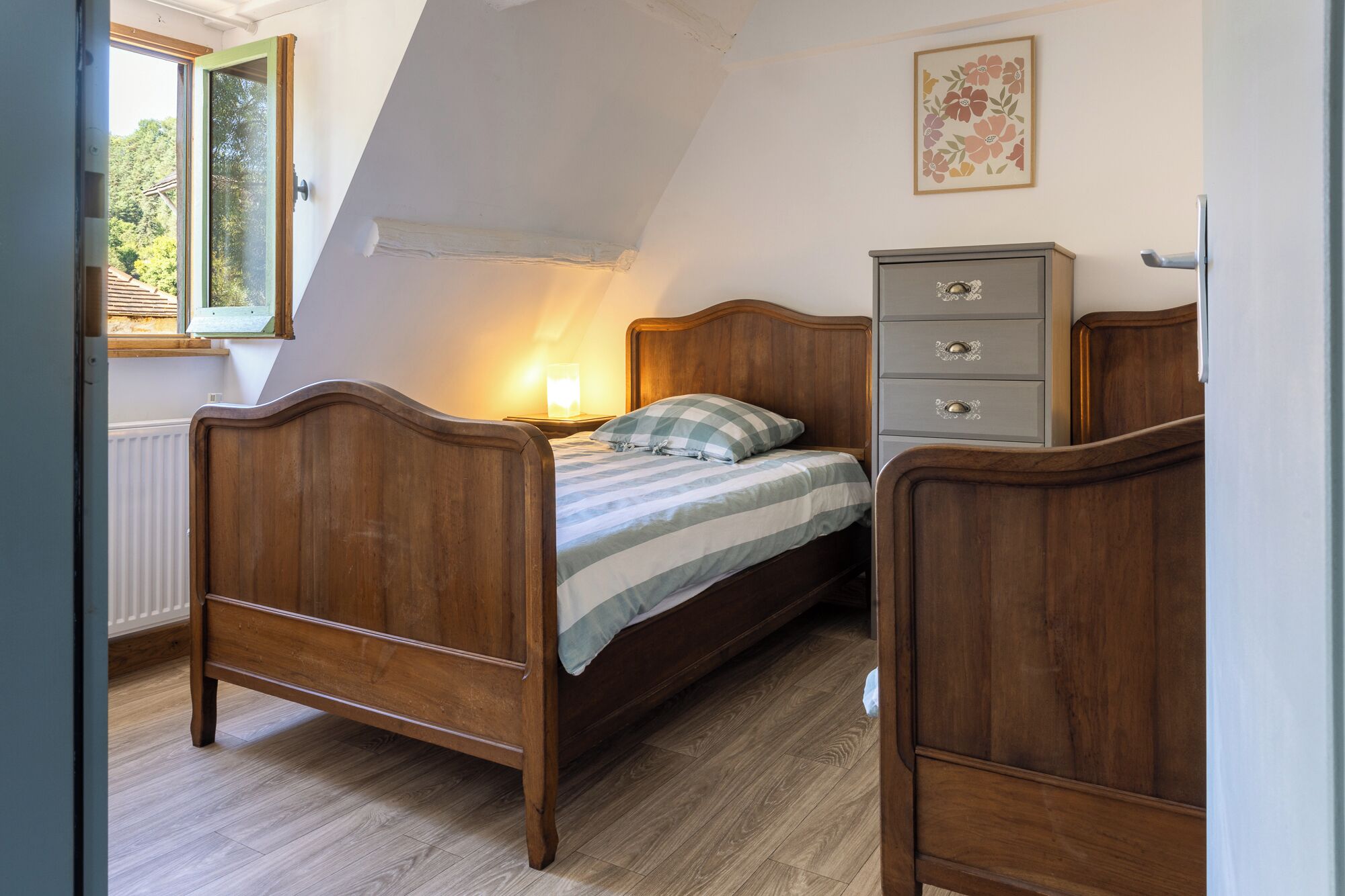 Un dormitorio con cama de madera, una mesita de noche con lámpara, un aparador y una ventana con vistas a árboles.