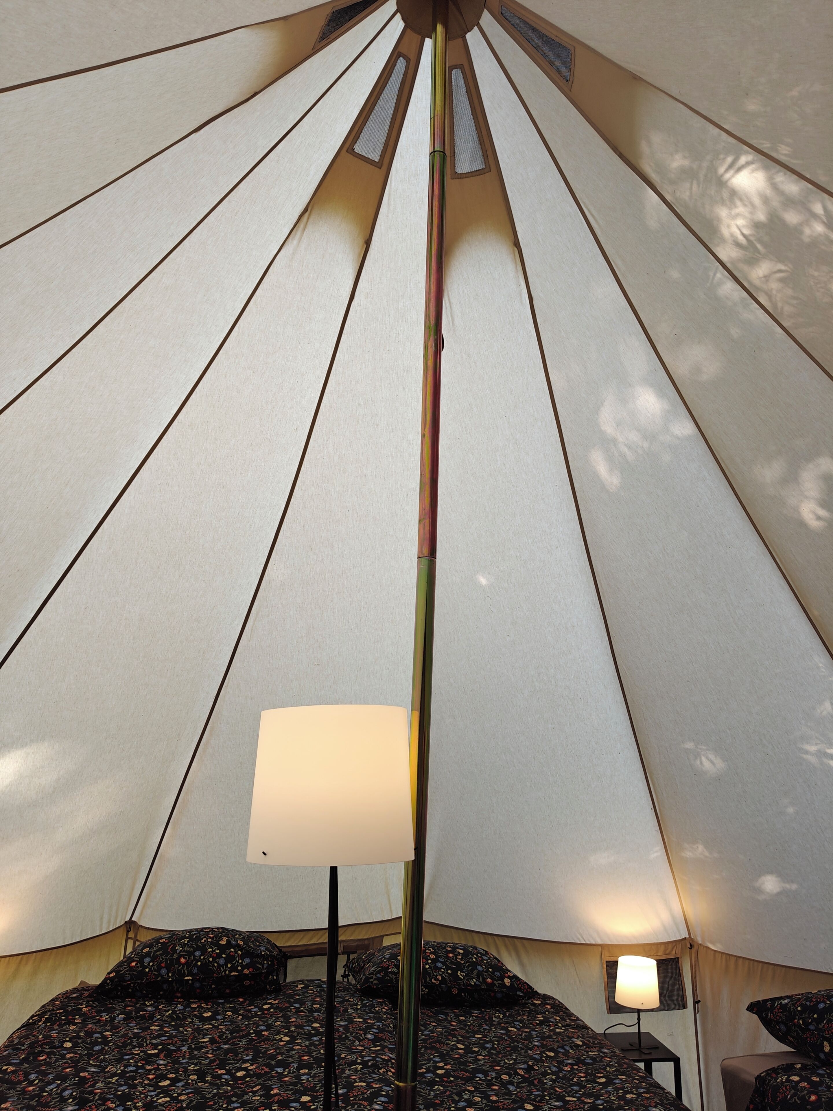 Ein Zeltinneres mit einer zentralen Stange, einem Bett mit blumigem Bettzeug und einer Tischlampe.