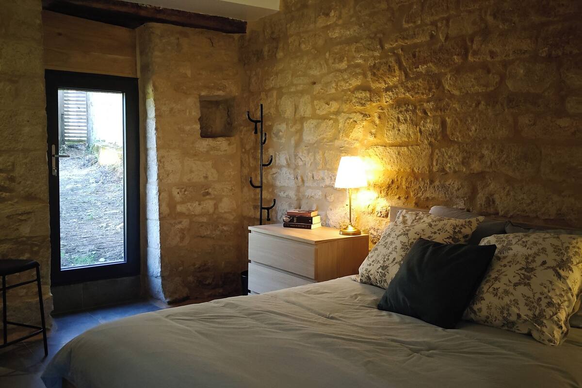 A bedroom with a stone wall, a bed with a white comforter, a nightstand with a lamp, and a window with a view outside.