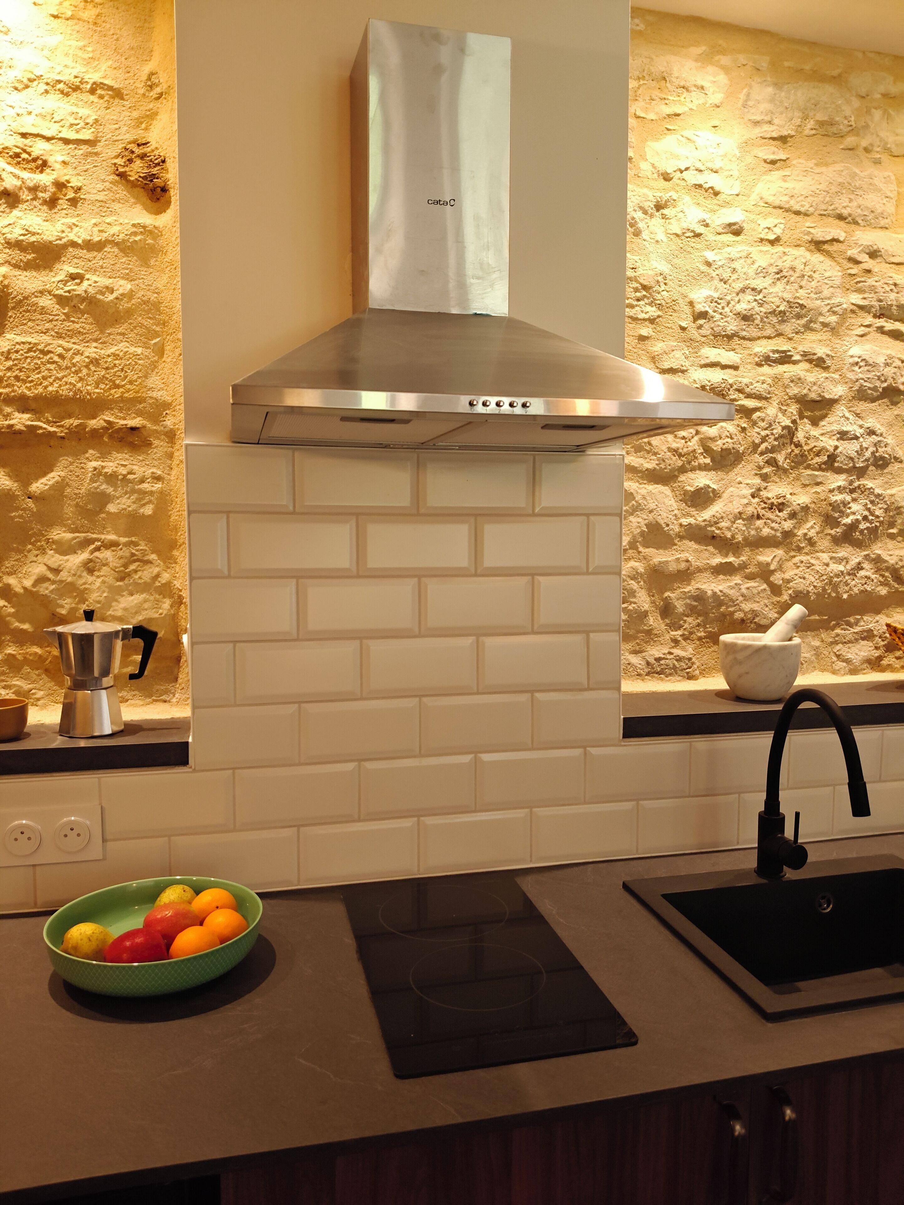 A kitchen with a stainless steel range hood, a stone-textured backsplash, a coffee maker, and a bowl of fruit on the counter.
