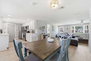 A modern kitchen with white cabinets, a wooden dining table, and a seating area with a sofa and chairs. - Liberty Breeze Retreat (Jacksonville Beach)