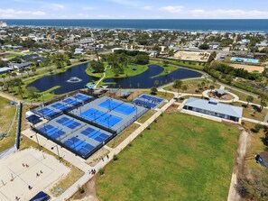 Vista aérea de un complejo deportivo con múltiples canchas de tenis, una piscina y un edificio con techo azul.