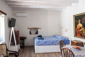 Een kamer met een bed, een bureau met een computer, een stoel, een tafel met fruit en een schilderij aan de muur.