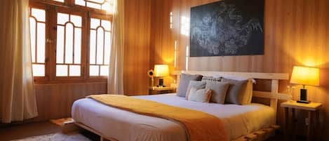 Una cama grande con cabecera, dos mesitas de noche con lámparas, un ventanal con marcos de madera y una obra de arte colgada en la pared.