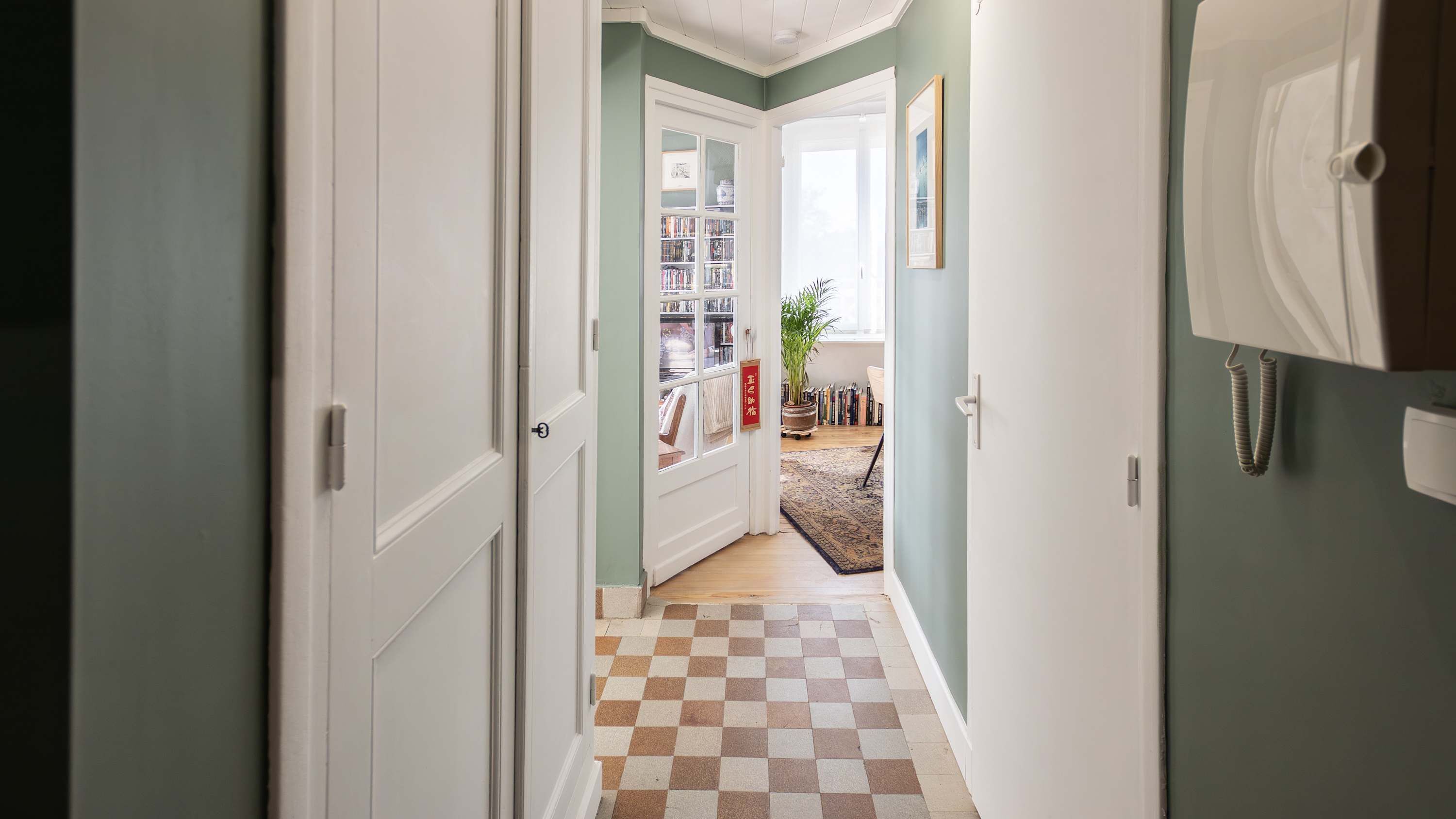Een hal met een zwart-wit geruit vloerkleed, een boekenkast en een deur naar een andere kamer.