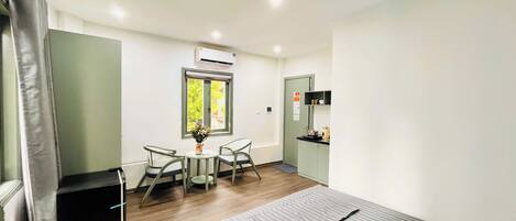 Una habitación pequeña con una cama, una mesa con sillas, un ventanal con vistas, un aire acondicionado y una kitchenette.