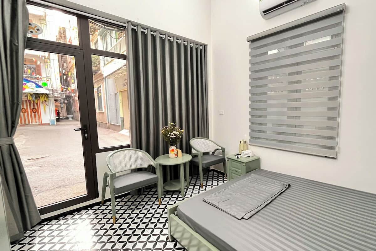 Una habitación pequeña y moderna con cama, una silla, una mesita con un jarrón y una ventana con persianas.