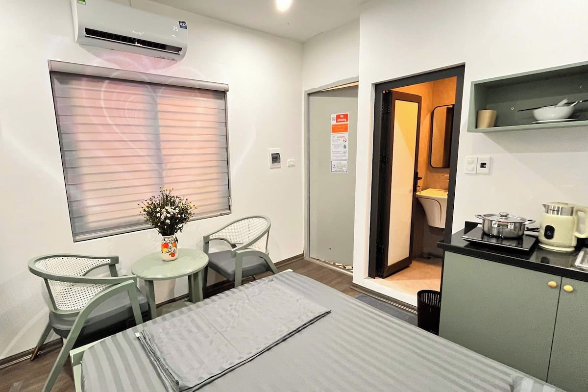 Una habitación de hotel compacta con cama, una pequeña mesa con un florero, dos sillas, una kitchenette y un baño visible a través de una puerta abierta.