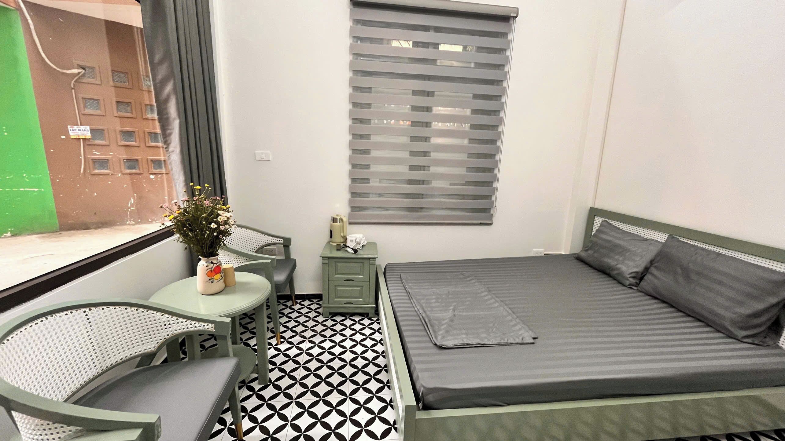 A small, neatly arranged room with a bed, a chair, a table, and a window with blinds.
