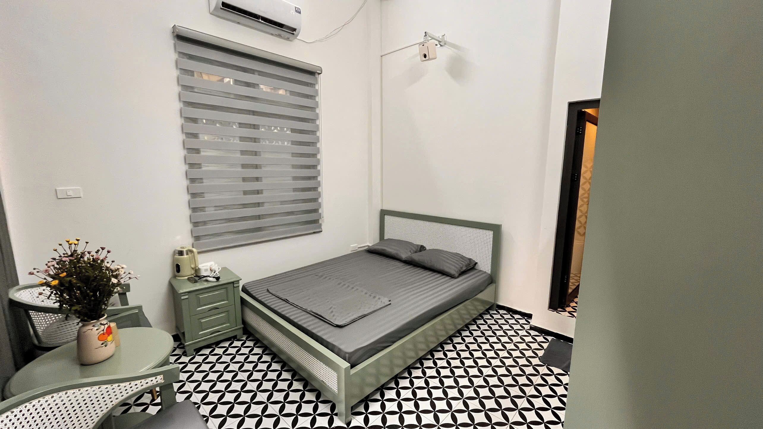 A small, clean room with a single bed, a bedside table, a chair, a small table with a vase, and a window with blinds.