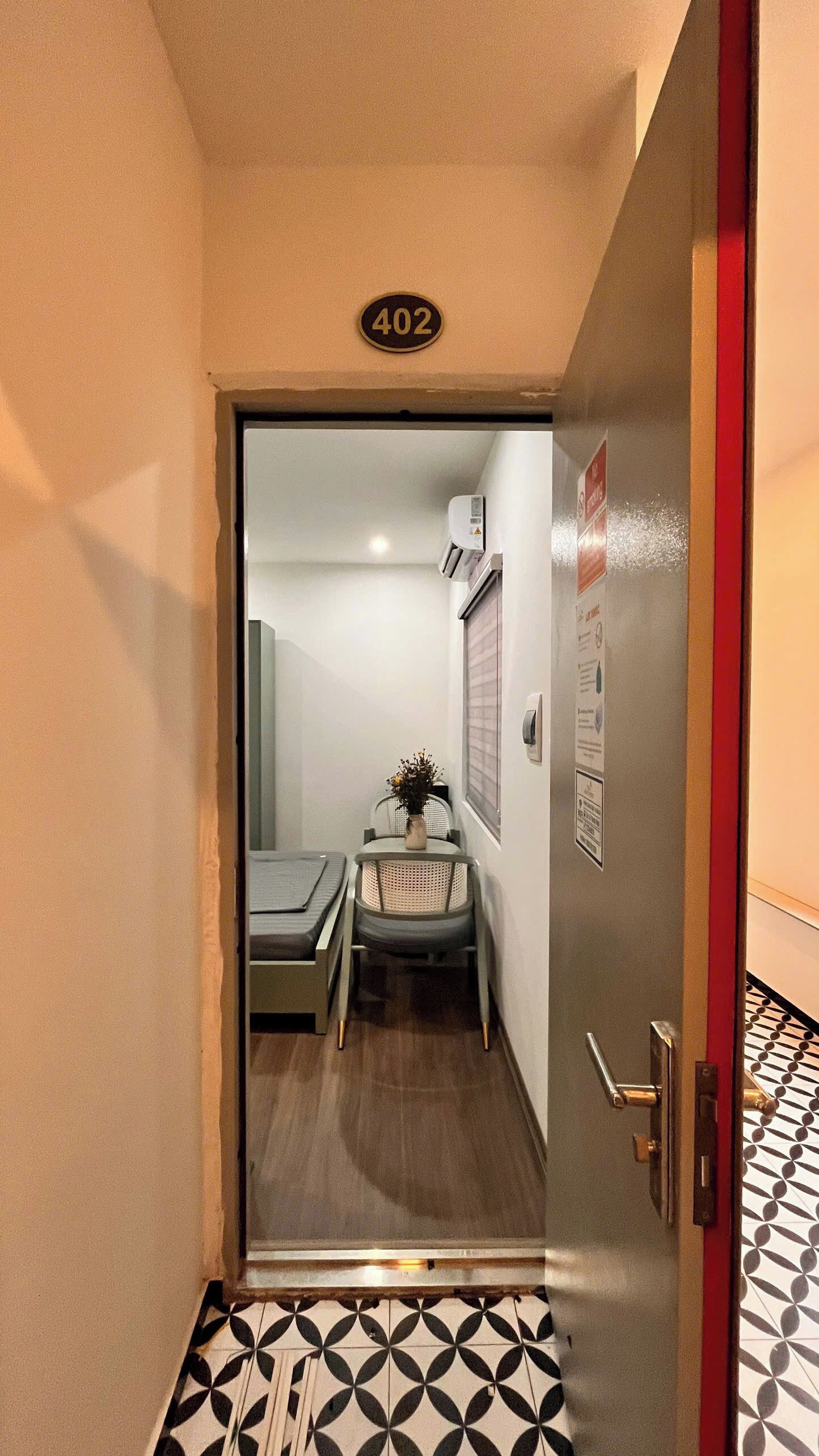 Een smalle gang met een deur waarop '402' staat, die leidt naar een kamer met een bed, een stoel en een raam.