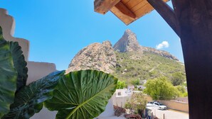 Vista de una montaña con un pico destacado, una hoja verde grande en primer plano y un edificio con fachada blanca al fondo.