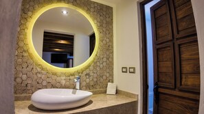 Ein Badezimmer mit rundem Spiegel, Waschbecken in Weiß, Steinwand und Holztür.