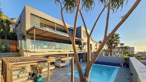 A modern two-story building with a rooftop pool, outdoor seating, and a tree in the foreground. - Room-luxury Couples Suite Ocean View & (Cape Town)