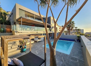 Un moderno edificio de dos pisos con piscina en la azotea, mobiliario exterior y un árbol en primer plano.