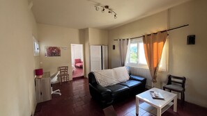 Un salon avec un canapé noir, une table basse blanche, une chaise en bois, un radiateur et une fenêtre avec des rideaux.