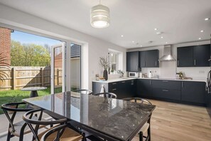 A modern kitchen with dark cabinets, a glass dining table, and a view of the backyard. - Premium Home, Garden & Parking, Nr Saunton Sands (Braunton)
