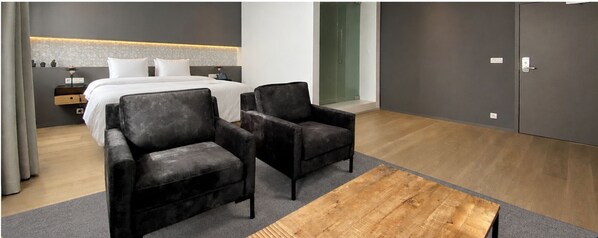 A modern hotel room with a bed, two armchairs, a wooden coffee table, a nightstand, and a wall-mounted lamp.