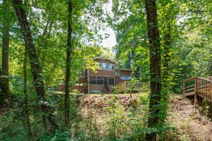 A two-story cabin surrounded by dense forest, with a wooden deck and a swing set. - Riverfront Cabin, Fenced Yard, Firepit & Grill (Lake Lure)