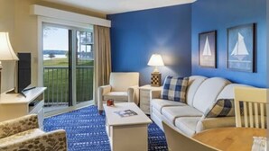 A hotel room with a sofa, armchair, coffee table, and a TV. There is a balcony with a view of the outdoors.