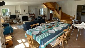 A dining area with a table set for four, surrounded by wicker chairs, in a house with a staircase and a living room in the background. - Belle-Île-en-Mer — Large family home 12 pers · Garden · 5 min from Donner (Bangor)