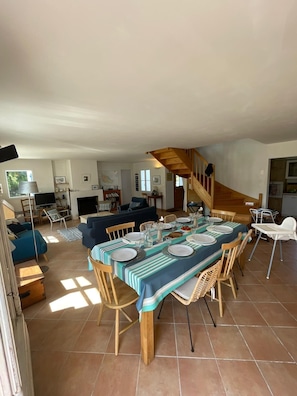 Ein Essbereich mit einem Tisch für vier Personen, umgeben von Korbstühlen, in einem Haus mit Treppe und Wohnzimmer im Hintergrund.