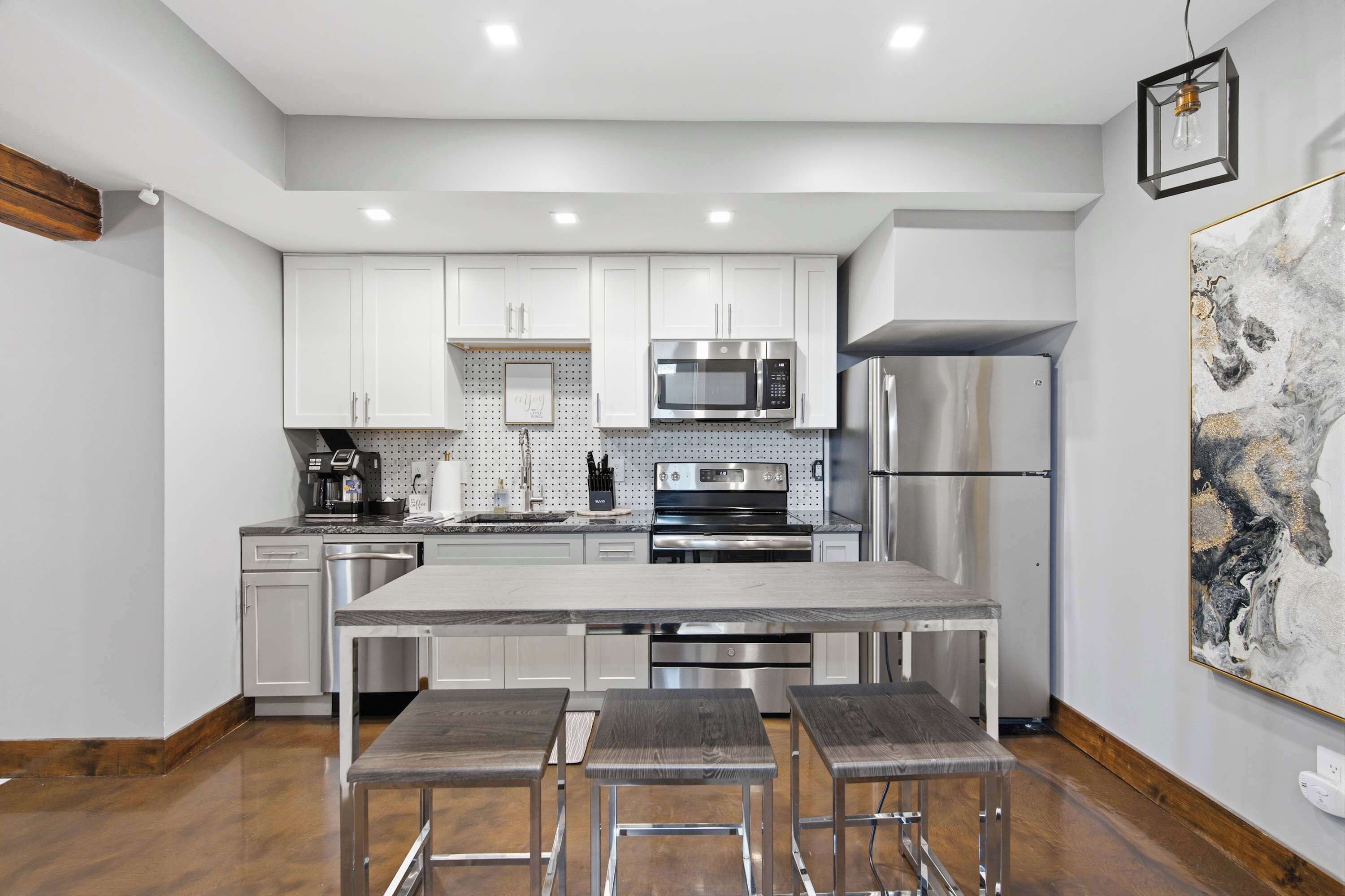 Cocina moderna con gabinetes blancos, electrodomésticos de acero inoxidable, encimera de mármol y una barra con taburetes.