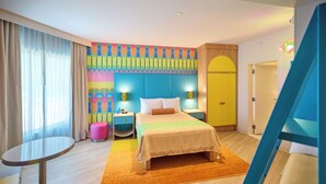 Una habitación de hotel con una cama, mesitas de noche, una mesa redonda, una pared colorida y una puerta.
